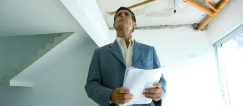 Homme en costume regardant un plafond avec des tuyaux exposés et tenant des plans. Inspection de travaux.