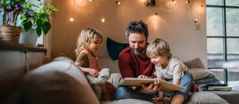 Père souriant lisant un livre à ses deux jeunes enfants sur un canapé, éclairés par des guirlandes lumineuses.