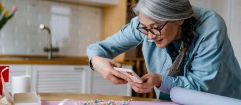 Une femme asiatique aux cheveux gris prend une photo de ses bijoux artisanaux avec un téléphone dans une cuisine.