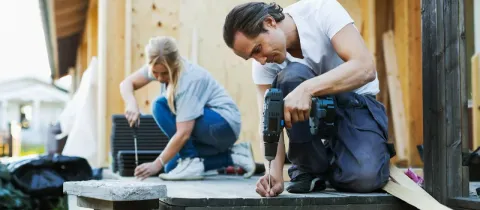 Un couple travaille ensemble pour construire une terrasse extérieure, utilisant des outils comme une visseuse et un tournevis.