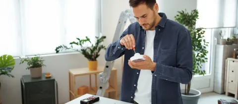 Un jeune homme répare ou installe un détecteur de fumée blanc sur une table remplie d'outils et d'appareils de mesure.