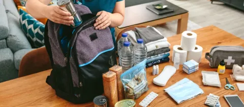 Préparation d'un sac d'urgence avec de la nourriture en conserve, des bouteilles d'eau et des fournitures médicales sur une table.