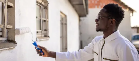Jeune homme souriant en combinaison blanche peignant un mur extérieur blanc près d'une fenêtre à l'aide d'un rouleau.