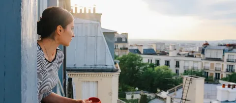 Femme buvant une boisson chaude, regardant le paysage urbain de Paris depuis un balcon ensoleillé.
