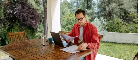 Homme en chemise rouge travaillant sur ordinateur portable et documents à une table en bois, en extérieur sous un auvent.