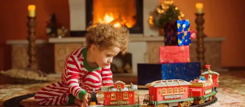 Petite fille en pyjama rayé jouant avec un train électrique de Noël devant une cheminée allumée.