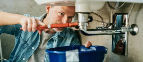 Plombier ou propriétaire réparant une fuite sous un lavabo avec une clé à molette, un seau en dessous.
