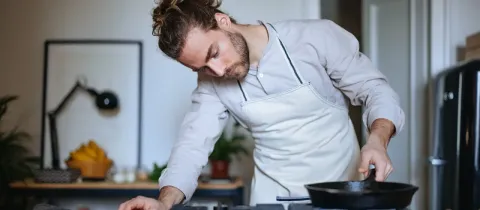 Un homme avec un chignon et un tablier se penche sur une cuisinière à gaz, concentré sur la poêle en fonte qu'il est en train de chauffer.