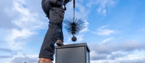 Un ramoneur sur un toit tient la brosse de ramonage au bout d'une chaîne, se préparant à nettoyer une cheminée.