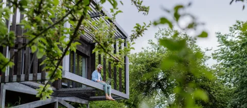 Un homme barbu travaille sur un ordinateur portable, assis sur le rebord d'un balcon en bois d'une maison moderne au milieu des arbres.