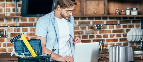 Un homme avec des lunettes utilise un ordinateur portable sur un plan de travail, à côté d'une boîte à outils ouverte, pour une réparation.
