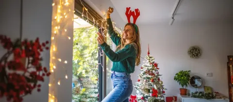 Une jeune femme joyeuse portant des bois de renne décore une fenêtre avec des guirlandes lumineuses à côté d'un arbre de Noël.