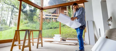 Un homme barbu en chemise à carreaux examine un plan dans une pièce en construction avec de grandes baies vitrées.