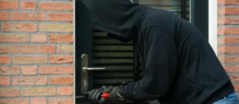 Un cambrioleur masqué, vêtu de noir, tente de forcer une serrure de porte avec un outil.