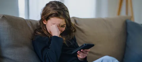 Jeune fille assise sur un canapé, l'air triste et se frottant les yeux en regardant son téléphone portable.