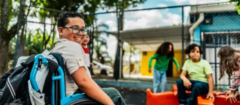 Un garçon souriant à lunettes en fauteuil roulant s'amuse avec ses camarades dans une cour de récréation.