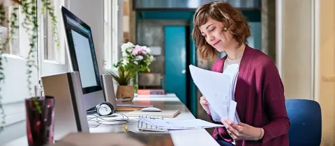 Une employée de bureau souriante assise à son poste de travail examine des documents administratifs papier.