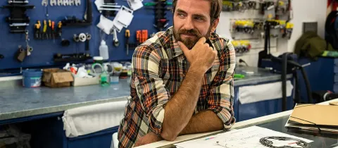 Un homme concentré, assis à une table dans un atelier, avec divers outils et équipements autour de lui.