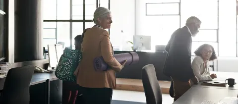 Une femme souriante traverse un bureau moderne en portant un tapis de yoga violet sous le bras.