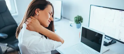 Une employée de bureau assise exprime une vive douleur au cou et aux cervicales devant son ordinateur portable.