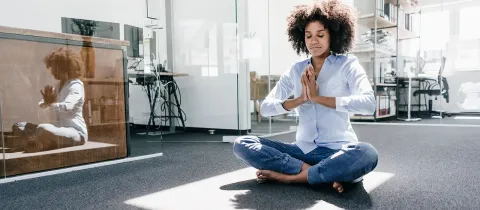 Une employée aux cheveux crépus médite assise en tailleur sur le sol de son bureau dans un rayon de soleil.