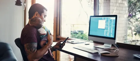 Homme en télétravail tenant son bébé dans les bras face à son ordinateur et son smartphone.