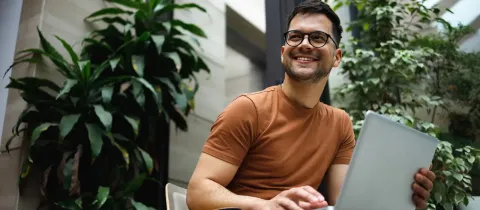 Un homme souriant, assis à l’extérieur entouré de plantes vertes, travaille sur un ordinateur portable.