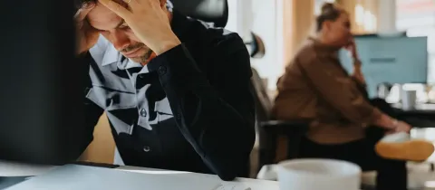 Un homme stressé, assis à un bureau, se tient la tête devant un ordinateur, dans un environnement de travail.