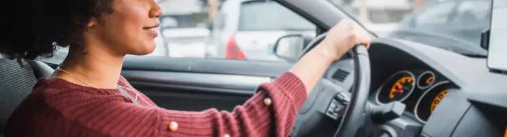 Une femme conduit une voiture en souriant, vue depuis l'intérieur du véhicule.