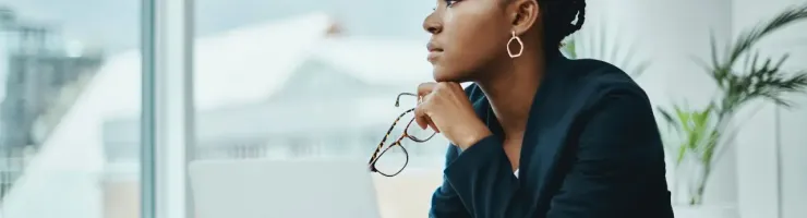Femme pensive regardant par la fenêtre avec un ordinateur devant elle.