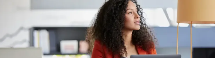 Femme aux cheveux bouclés assise à un bureau avec un ordinateur portable, regardant pensivement sur le côté.