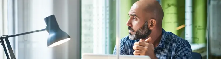 Homme chauve avec barbe assis à un bureau avec un ordinateur portable, regardant sur le côté.