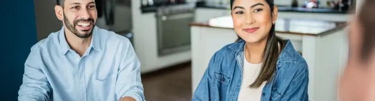 Couple souriant assis à table dans une cuisine lumineuse, regardant quelqu'un hors cadre.