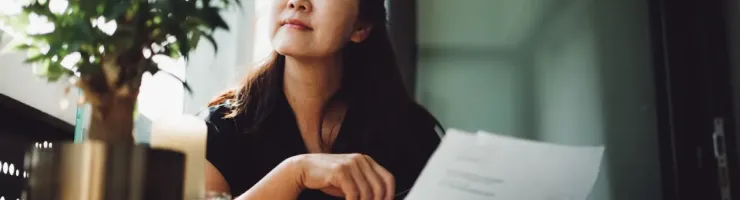 Femme tenant des documents, regardant vers le haut et réfléchissant, près d'une fenêtre avec des plantes.