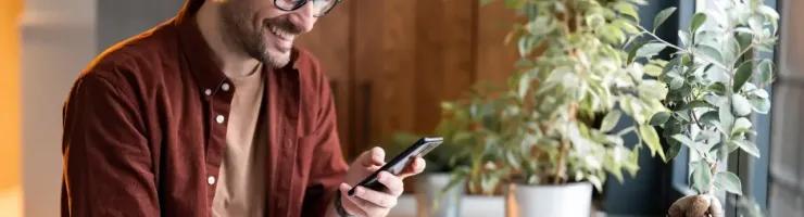 Homme souriant avec lunettes, utilisant téléphone et ordinateur portable près d'une fenêtre avec plantes.