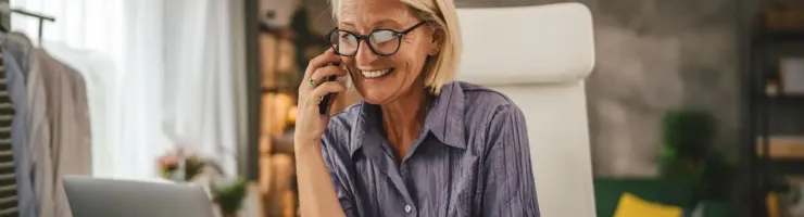 Femme agée souriante avec des lunettes parle au téléphone en travaillant sur un ordinateur .