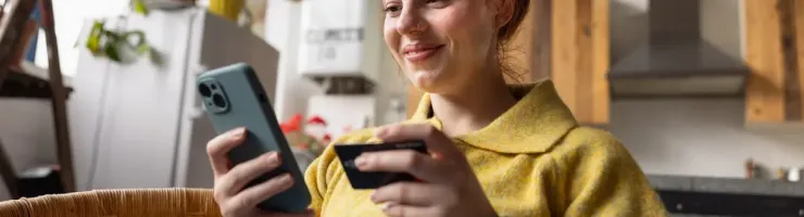 Femme souriante assise dans une chaise, tenant un téléphone et une carte bancaire.