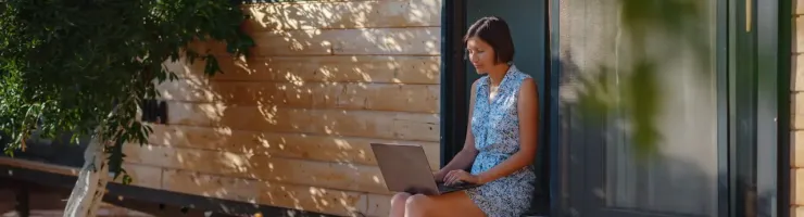 Femme assise sur les marches d'une tiny house en bois, sur un ordinateur portable.