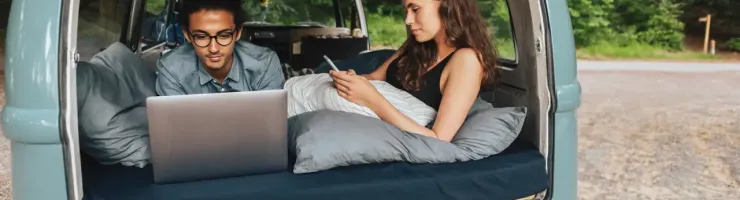 Couple assis à l'arrière d'un van aménagé, utilisant un ordinateur portable et un téléphone.