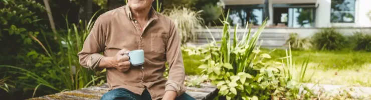 Homme d'âge mûr assis pieds nus sur une jetée en bois, tenant une tasse.