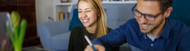 Un homme écrit dans un carnet, une femme souriante regarde un ordinateur portable à côté de lui.