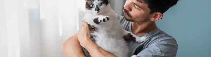 Un homme tenant un chat aux marques sombres et blanches près d'une fenêtre lumineuse.