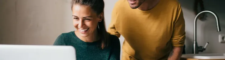 Un couple souriant regarde ensemble un ordinateur portable posé sur une table de cuisine.