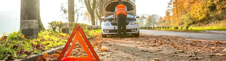 Un homme en gilet de sécurité orange examine le moteur d'une voiture en panne, avec un triangle de signalisation.