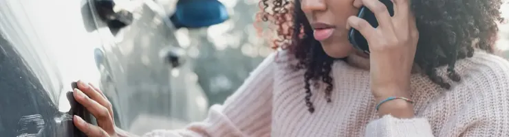 Une femme avec des cheveux bouclés, au téléphone, touche une éraflure sur le côté d'une voiture.