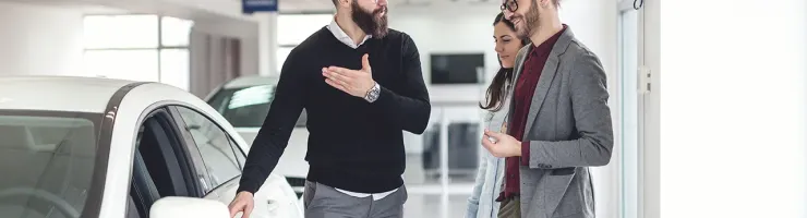 Un vendeur barbu discute avec un couple souriant dans un showroom de voitures, pointant vers une voiture blanche.