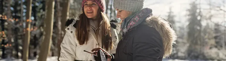 Deux femmes en vêtements d'hiver regardent un téléphone dans une forêt enneigée.