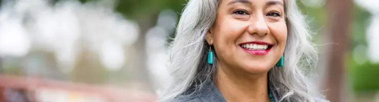Une femme souriante aux cheveux gris avec des boucles d'oreilles et un collier turquoise, sur fond de verdure.