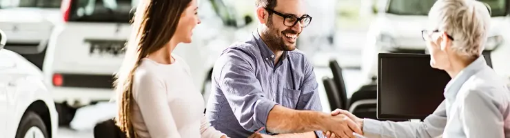 Un couple sourit et un homme serre la main d'une femme à un bureau dans un concessionnaire automobile.