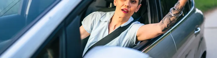 Une femme aux cheveux courts et tatouée lève la main en l'air depuis sa voiture, l'air exaspéré.